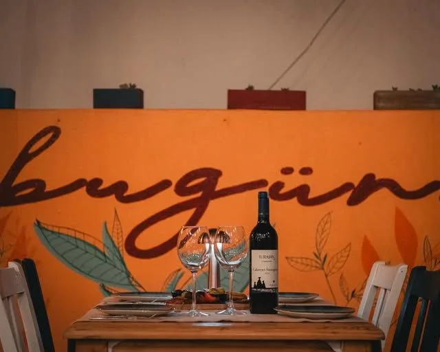 Turuncu duvarda el yazısı “bugün” logosu; önünde bir şişe şarap ve iki kadeh olan masa